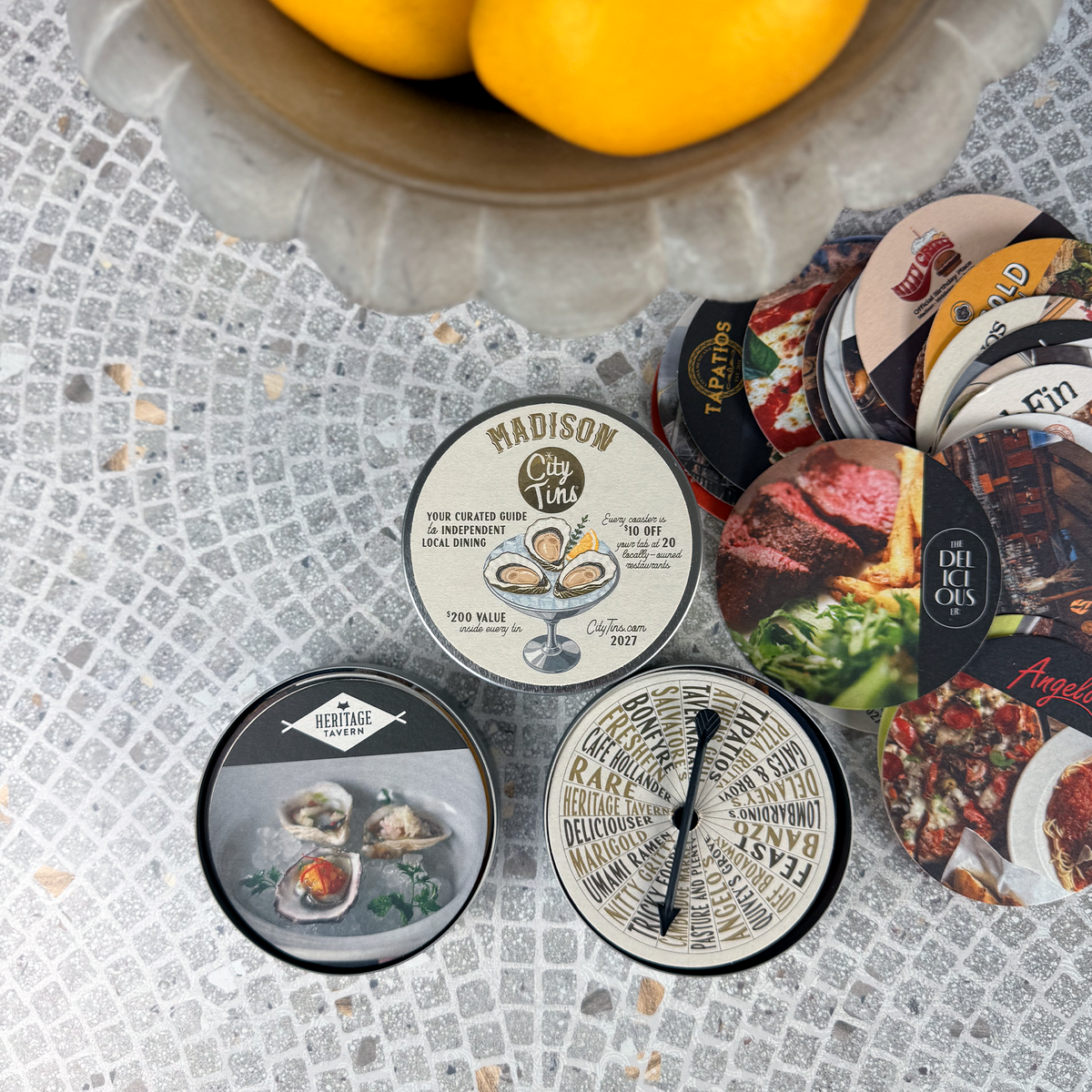 A styled flat-lay of coaster gift cards, featuring a central round tin labeled 'CityTins Madison' surrounded by tin contents of coasters featuring independent restaurants, with a bowl of lemons partially visible at the top on a gray tiled surface.