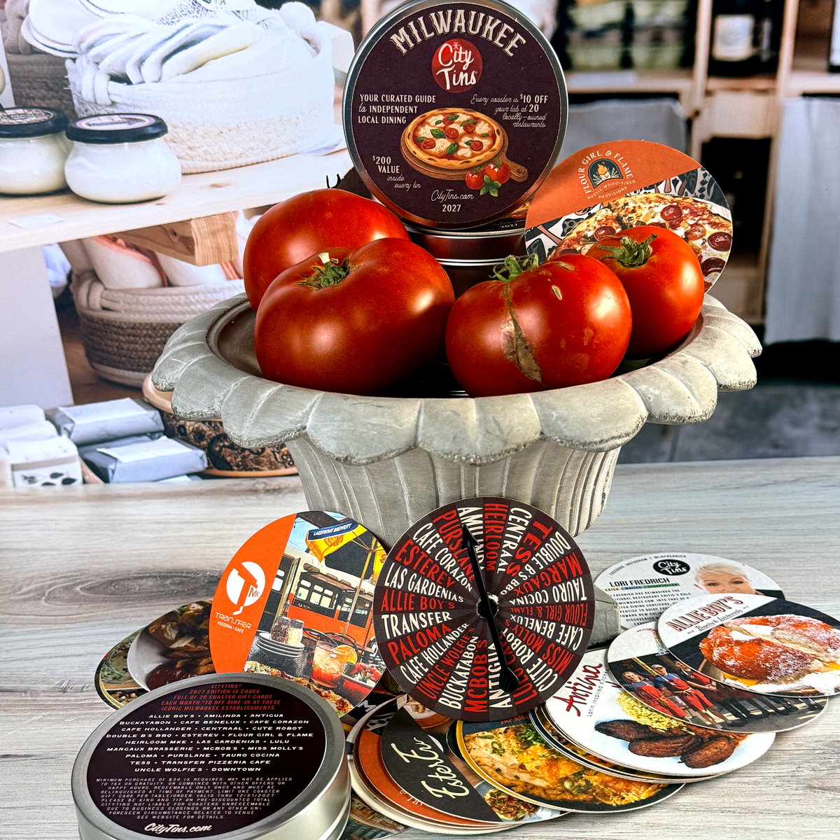 A collection of printed coaster gift cards featuring all independent restaurants in Metro-Milwaukee, including Four Girl & Flame, Antigua, Ester Ev and Allie Boys, displayed in a decorative stone bowl with fresh tomatoes on a wooden table.