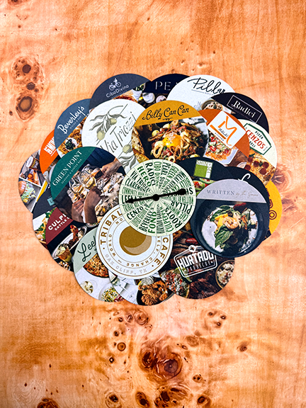 CityTins coaster gift cards displays on a wood table with the DinnerSpinner on top set to Leela's restaurant. Gift cards shown include Billy Can Can, Radici, Via Triozzi, Greenpoint, Hurtado and Tribal All Day Cafe.