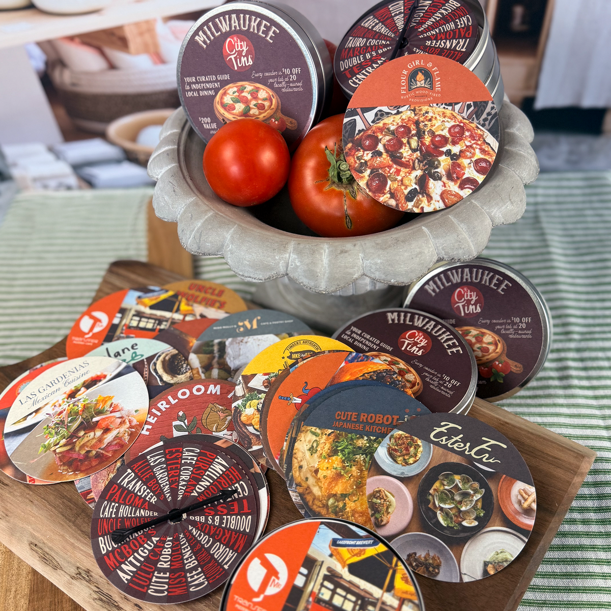 The 2027 collection of Milwaukee CityTins with a Dinner Spinner game piece and coasters featuring Cute Robot, Ester Ev, Las Gardenias and Heirloom, with a gray bowl holding two tomatoes in the background.