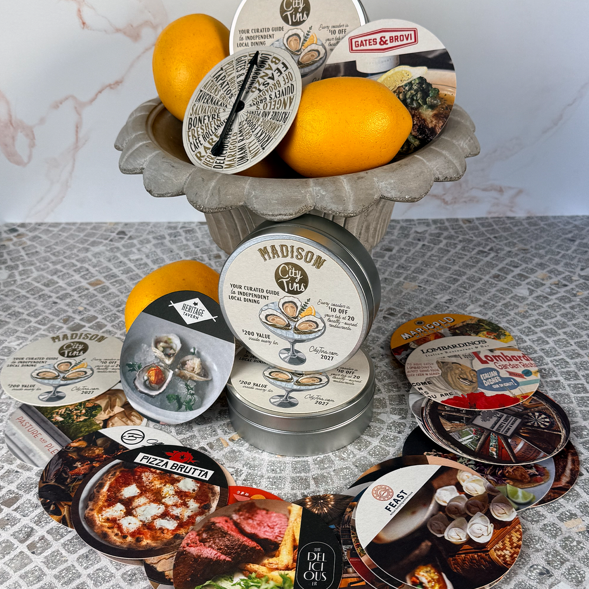 A collection of coaster gift cards arranged around a stone pedestal bowl filled with lemons. The product includes discounts at local restaurants shuch as Pizza Brutta, Heritage Tavern and The Deliciouser.
